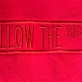 A close-up of bright red fabric with a vertical embroidered text reading 'FOLLOW THE SIGNS' in matching red thread.