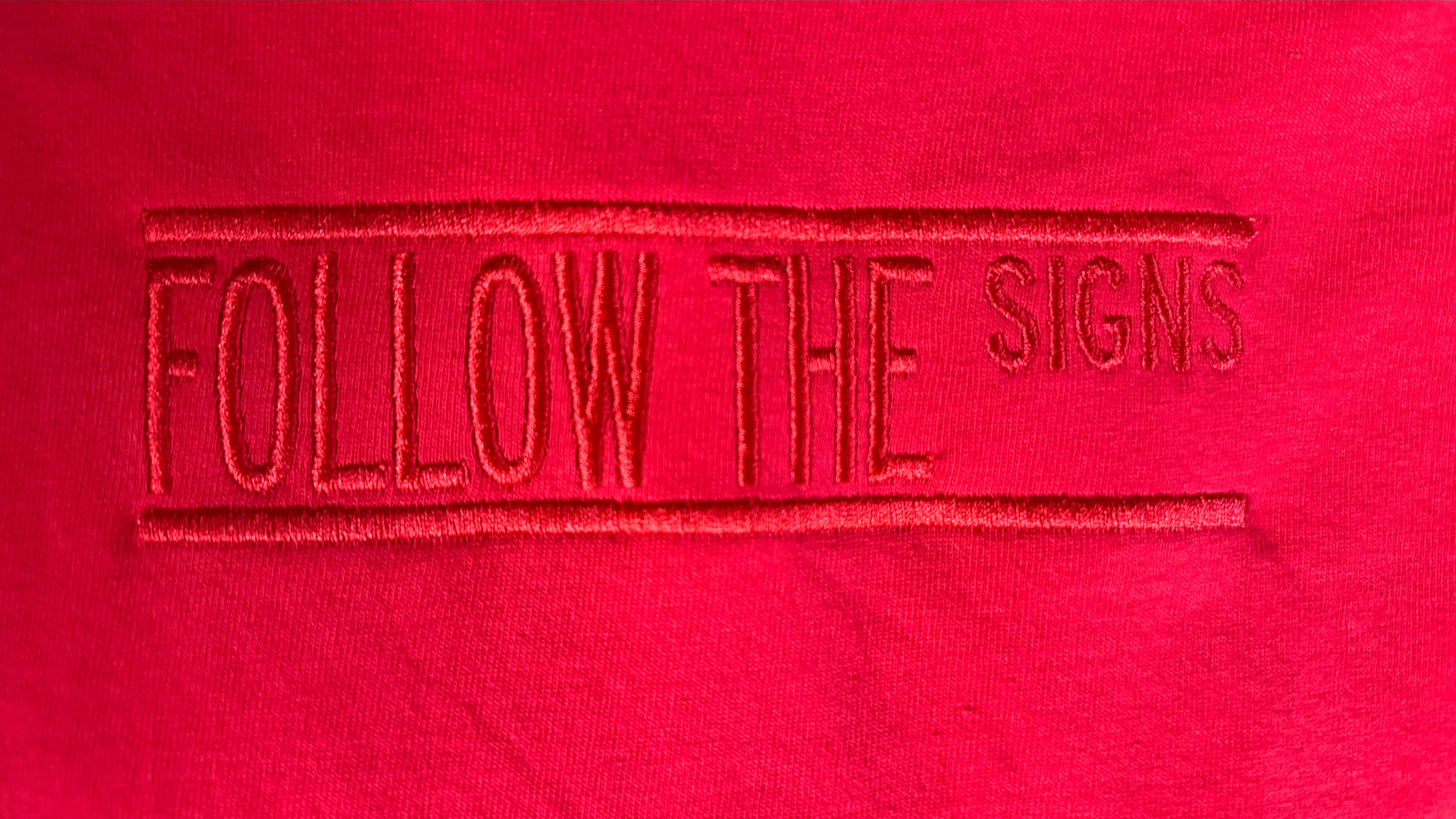 A close-up of bright red fabric with a vertical embroidered text reading 'FOLLOW THE SIGNS' in matching red thread.