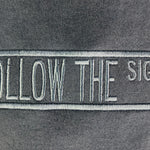 Close-up of a dark gray fabric surface with a vertical embroidered patch reading 'FOLLOW THE SIGNS' in light gray/white thread.