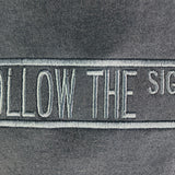 Close-up of a dark gray fabric surface with a vertical embroidered patch reading 'FOLLOW THE SIGNS' in light gray/white thread.