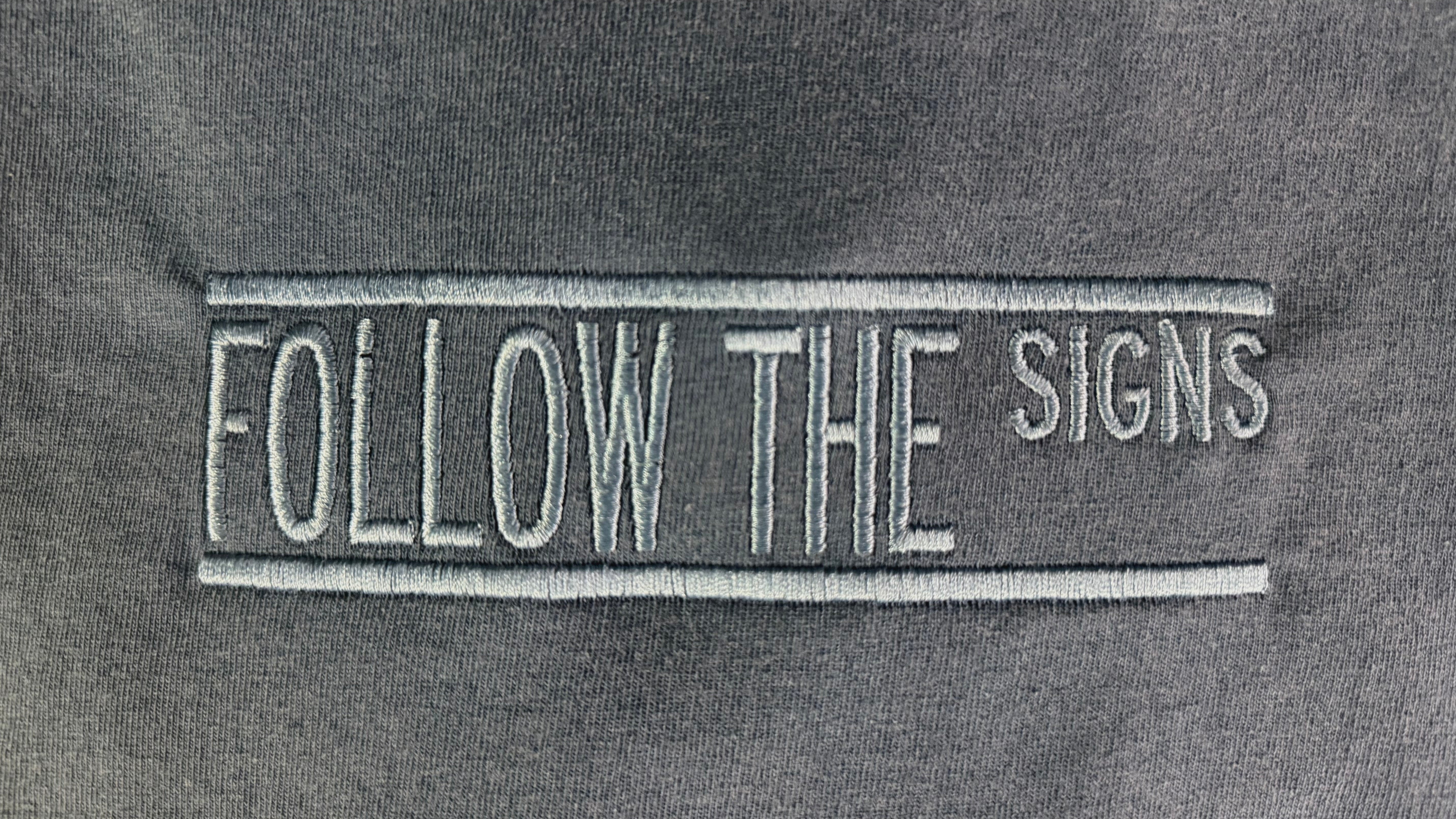Close-up of a dark gray fabric surface with a vertical embroidered patch reading 'FOLLOW THE SIGNS' in light gray/white thread.