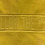 A close-up of a yellow fabric surface with vertical embroidered text reading 'FOLLOW THE SIGNS' in a matching yellow thread.