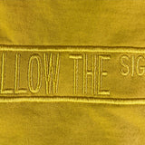 A close-up of a yellow fabric surface with vertical embroidered text reading 'FOLLOW THE SIGNS' in a matching yellow thread.