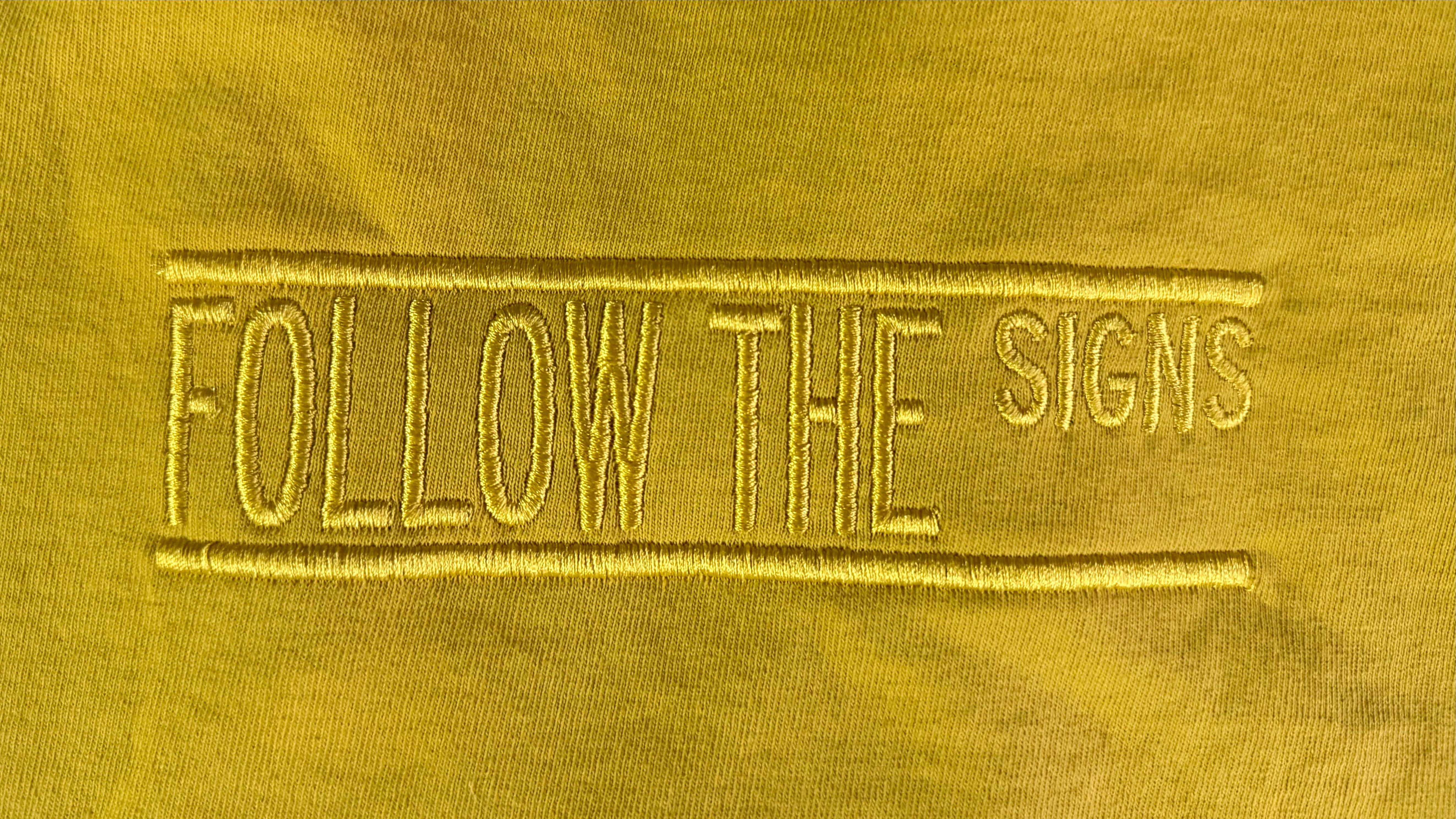 A close-up of a yellow fabric surface with vertical embroidered text reading 'FOLLOW THE SIGNS' in a matching yellow thread.