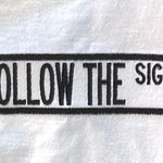 A close-up of a white fabric item, likely a towel or bandana, featuring a black embroidered vertical design that reads 'FOLLOW THE SIGNS' inside double border lines.