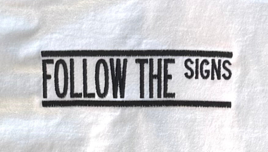 A close-up of a white fabric item, likely a towel or bandana, featuring a black embroidered vertical design that reads 'FOLLOW THE SIGNS' inside double border lines.