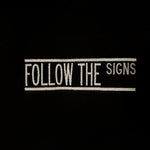A close-up of a black fabric surface featuring a vertical white embroidered text design that reads 'FOLLOW THE SIGNS' between two vertical lines.