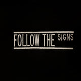 A close-up of a black fabric surface featuring a vertical white embroidered text design that reads 'FOLLOW THE SIGNS' between two vertical lines.