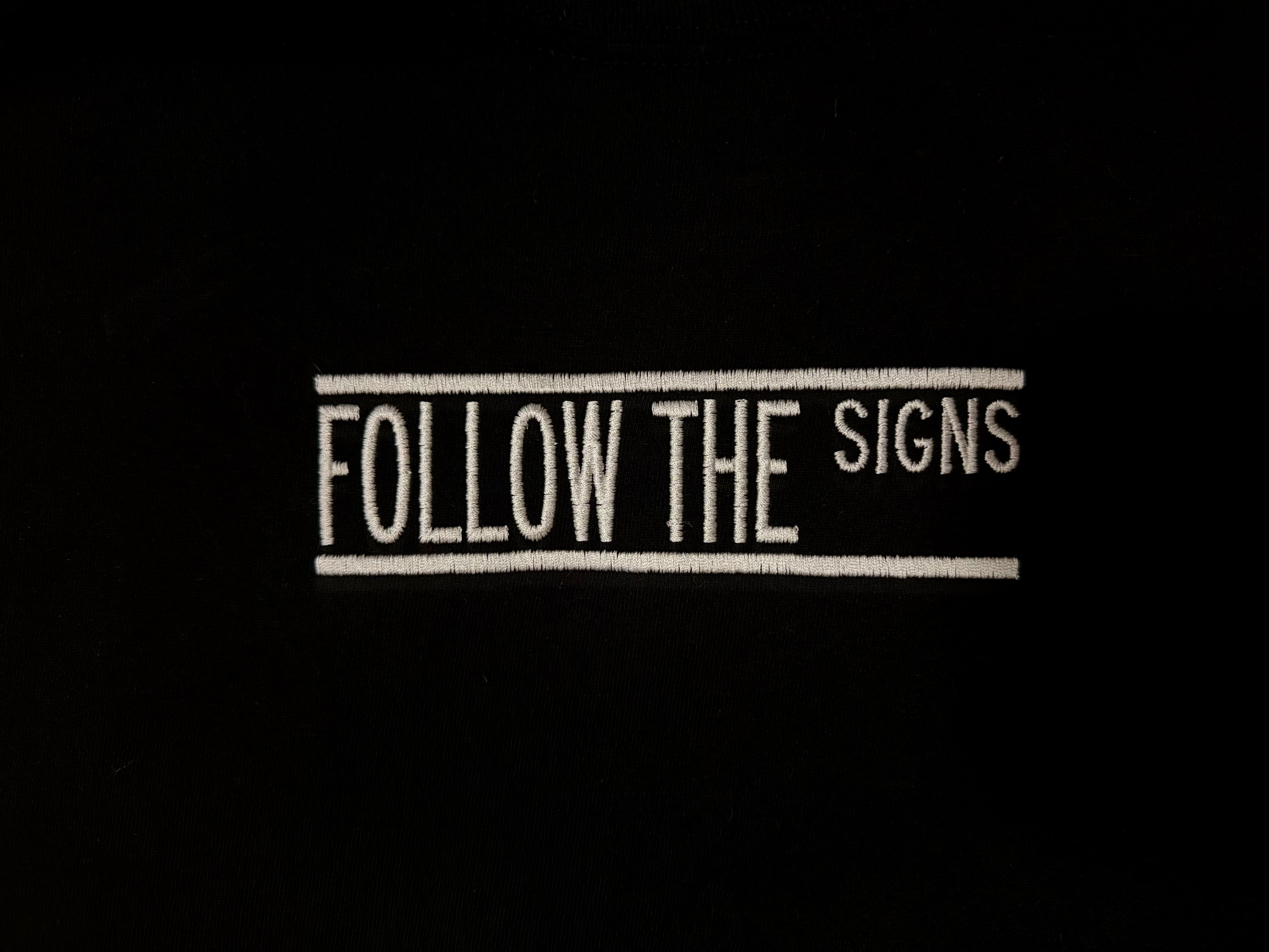 A close-up of a black fabric surface featuring a vertical white embroidered text design that reads 'FOLLOW THE SIGNS' between two vertical lines.